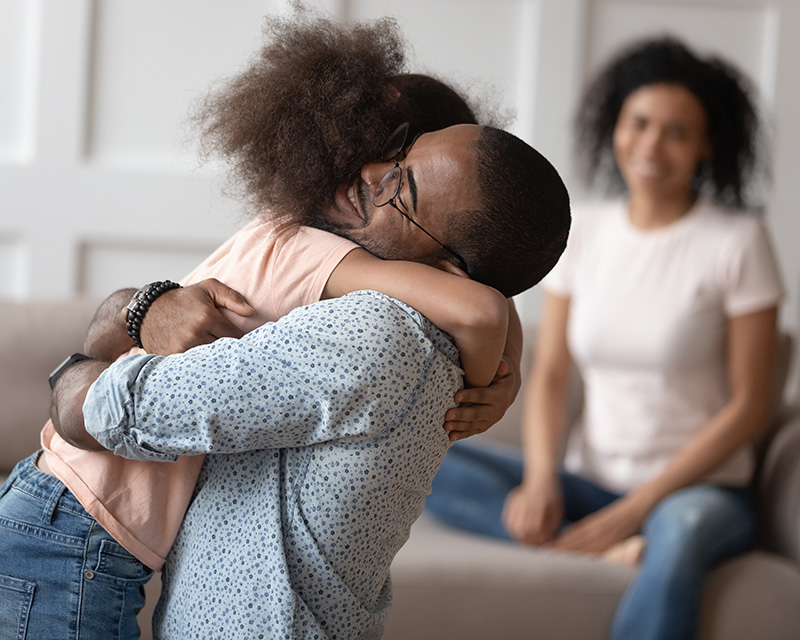 Happy little biracial girl hug loving african American dad meeting him at home after work business trip, overjoyed mixed race kid embrace black father reunited with smiling mom. Family reunion concept
